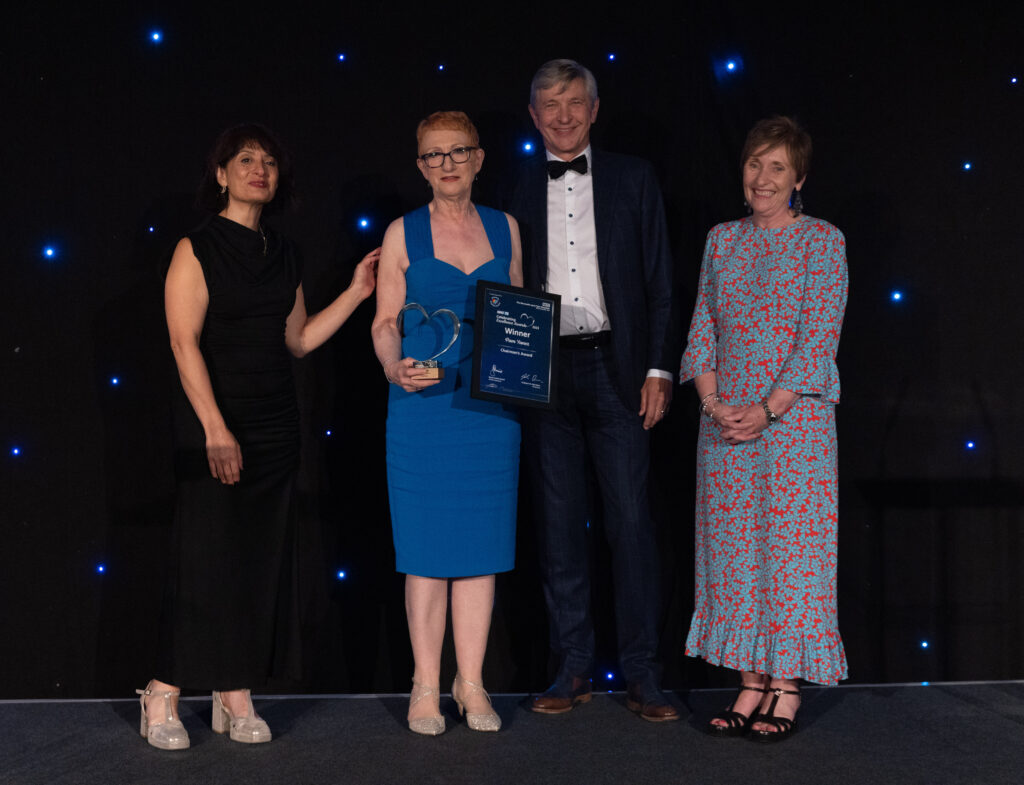 Picture of Pam Yanez stood on stage posing for a photo holding the award and certificate in front of star backdrop, with comedian Shaparak Khorsandi, Chairman Professor Sir John Burn and Maurya Cushlow.