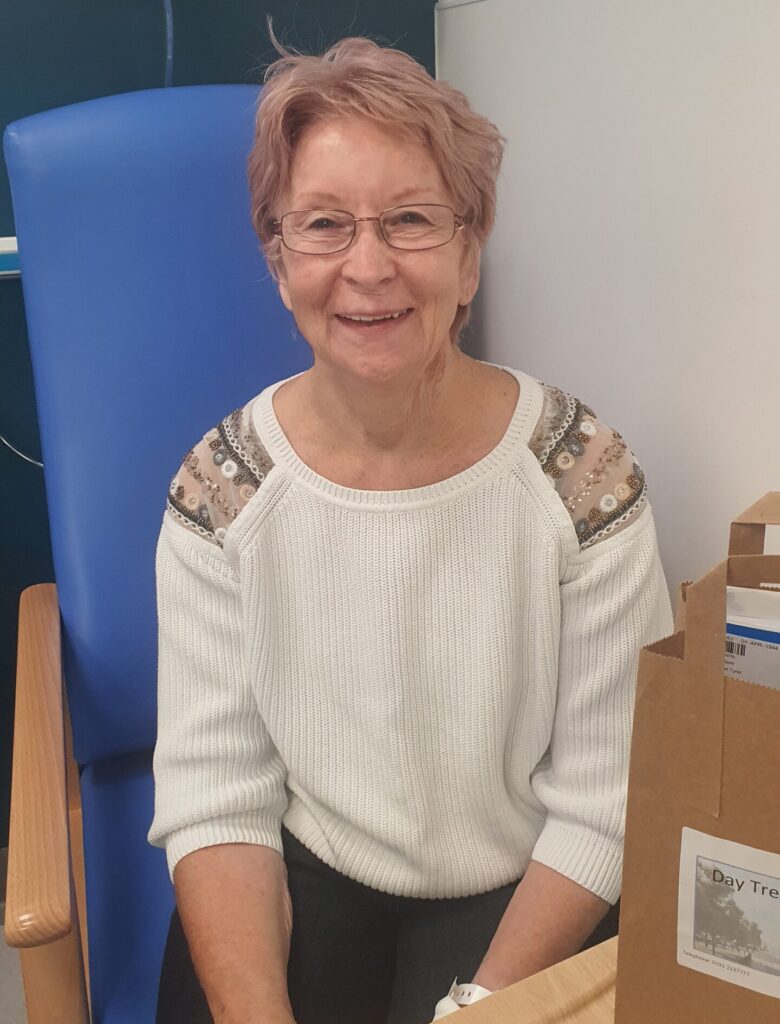 Photo shows lady with short fair hair, and glasses sitting in a hospital chair. She is wearing a cream jumper.