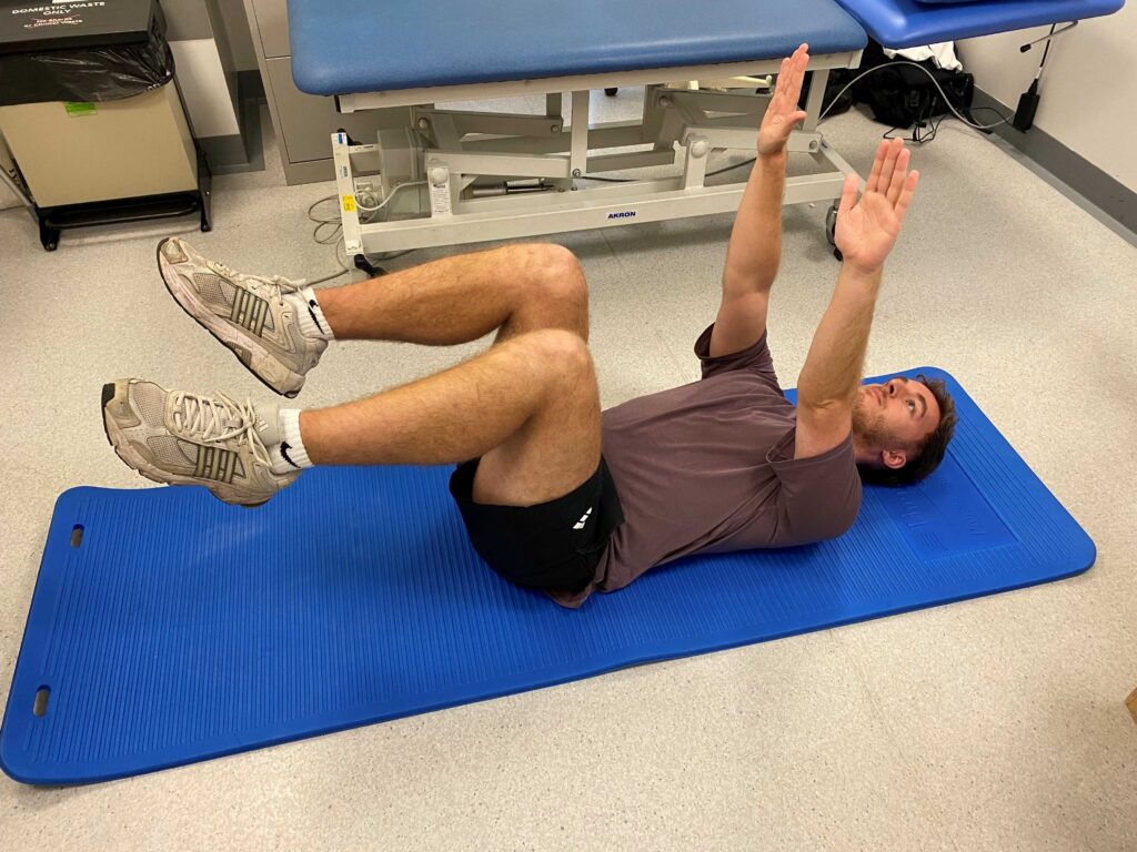Picture of someone lying on a blue mat on their back with hips and knees bent to 90 degrees and arms out straight with your fingertips pointing to the ceiling.