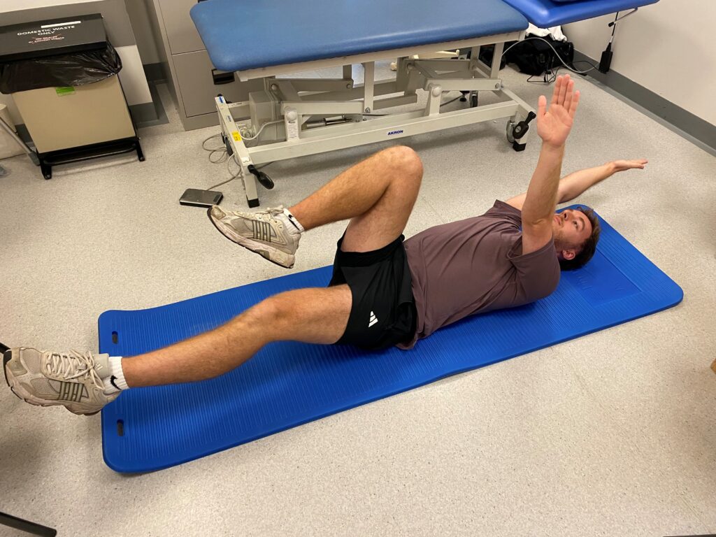 Picture of someone lying on a blue mat on their back with hips and knees bent. Keeping your back flat, lower your left arm and right leg away from one another towards the floor.