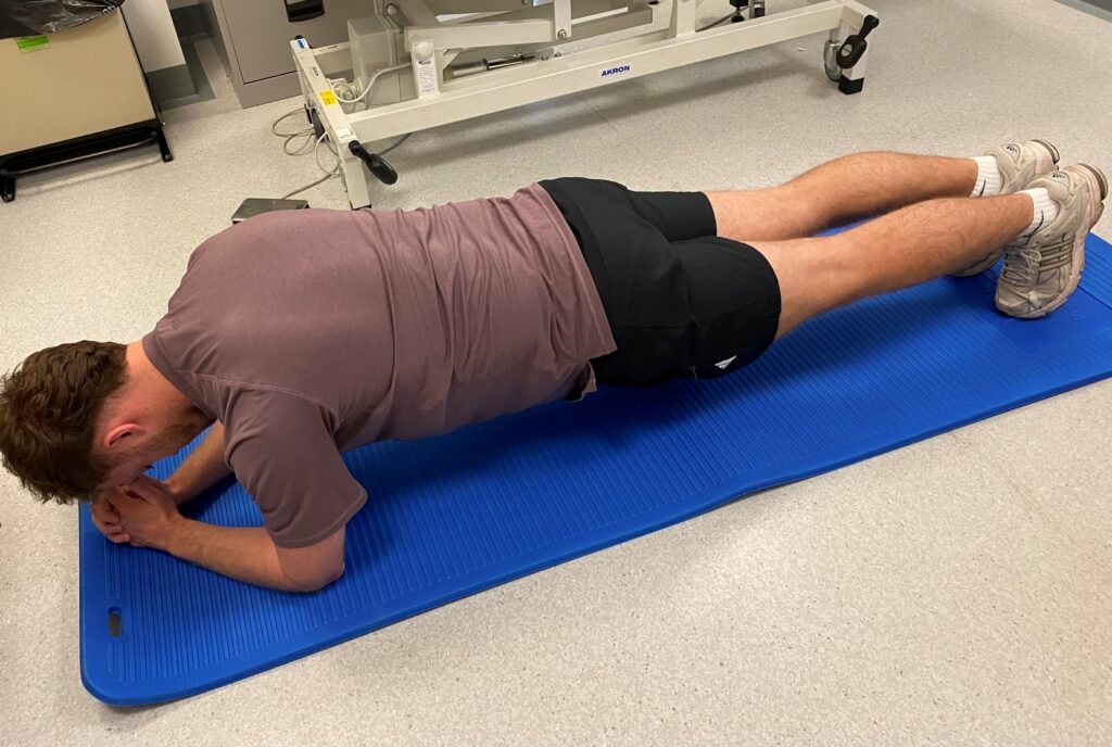 Picture of someone on a blue mat on the floor, rest on your forearms and toes with your body in a neutral alignment. Hold for 15-60 secs. Repeat 2-3 times.