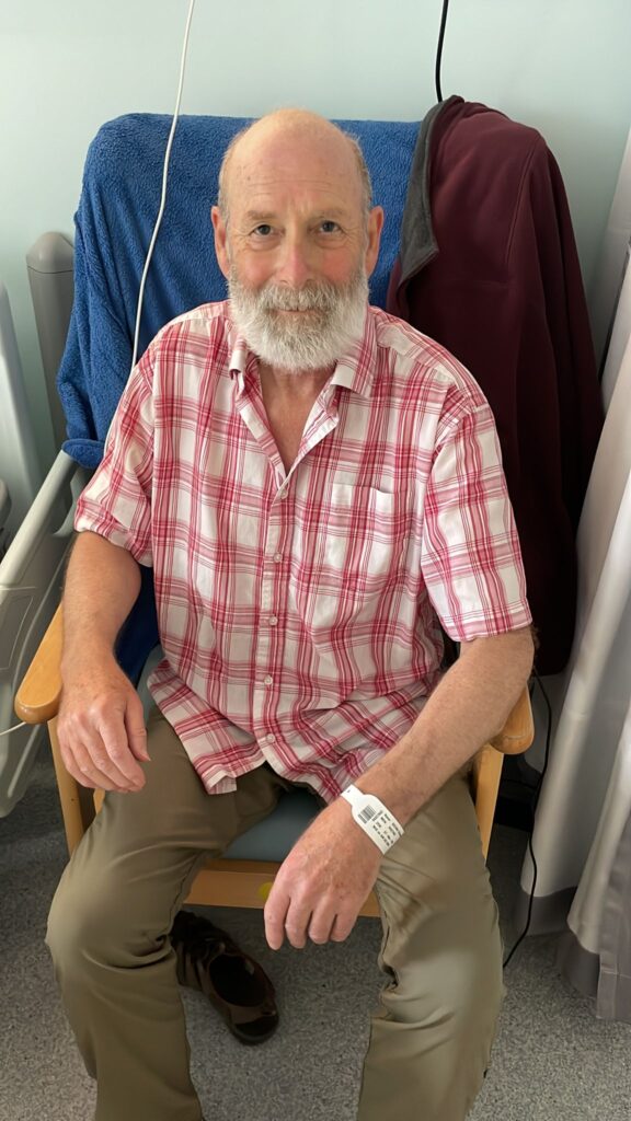 Chris is pictured sat in a hospital chair on a ward