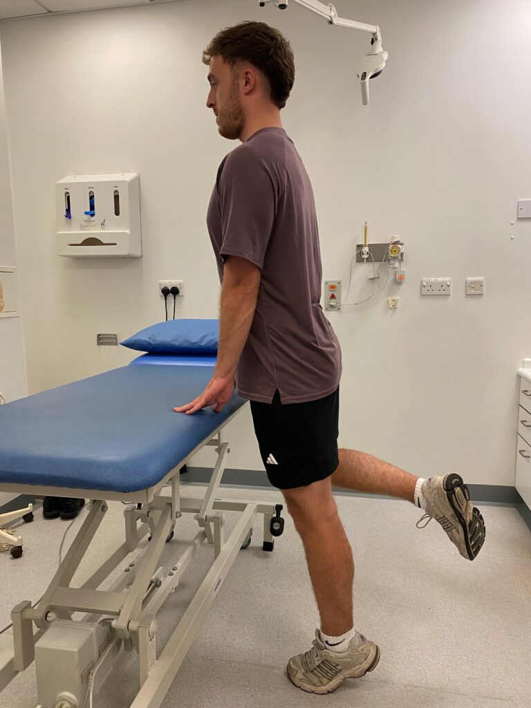 Person stood on one leg, with one leg bent holding onto a bed. 