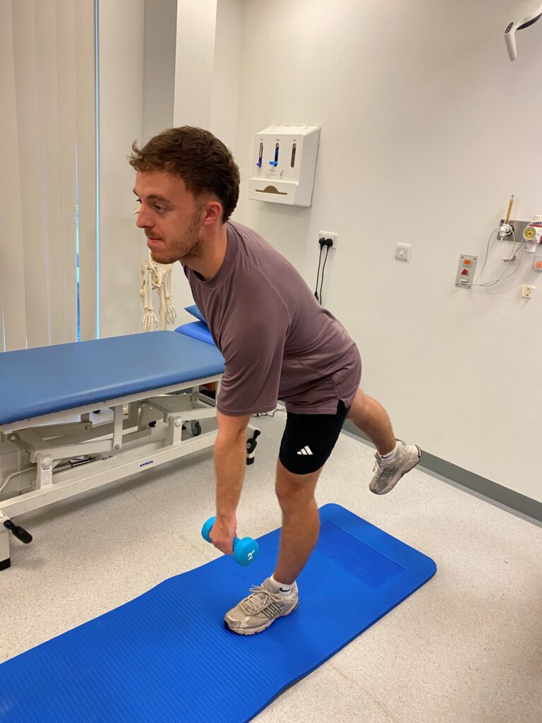 Person stood on a blue mat with knees slightly bent, holding a dumbbell in one hand.