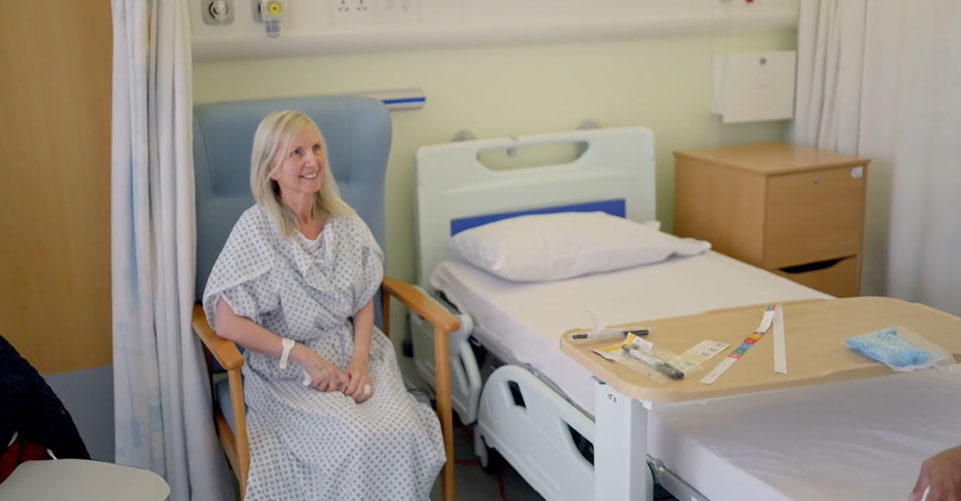 Picture of Susan sat on blue chair in surgical gown on the ward.